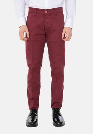 Uomo con pantaloni slim fit bordeaux, scarpe eleganti nere lucide e camicia bianca a maniche lunghe, in piedi davanti a uno sfondo semplice.