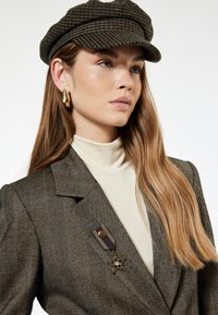 Bruine en zwarte visgraatblazer met een gouden broche, beige coltrui en een donkere, gestructureerde pet. Lang, steil haar.