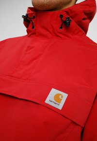 Red waterproof jacket with high collar, adjustable drawstrings, and a prominent yellow Carhartt logo patch on a white background.