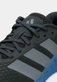 Μαύρο αθλητικό παπούτσι με γκρι επάνω μέρος από δίχτυ και τρεις ασημένιες ρίγες. Διαθέτει υφή υφάσματος, στρογγυλεμένο δάχτυλο και μπλε λαστιχένια σόλα.