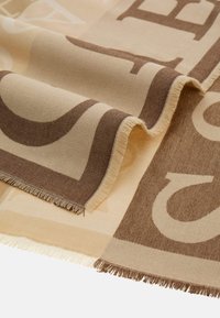 Coperta in tessuto intrecciato di colore crema e marrone, con grandi lettere grafiche. I bordi presentano dettagli con frangia, evidenziando una texture morbida.