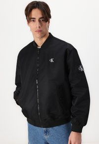 Čierna bomber bunda so zipsom, rebrovanými manžetami a golierom. Obsahuje vyšívané logo na hrudi a nášivku na rukáve.