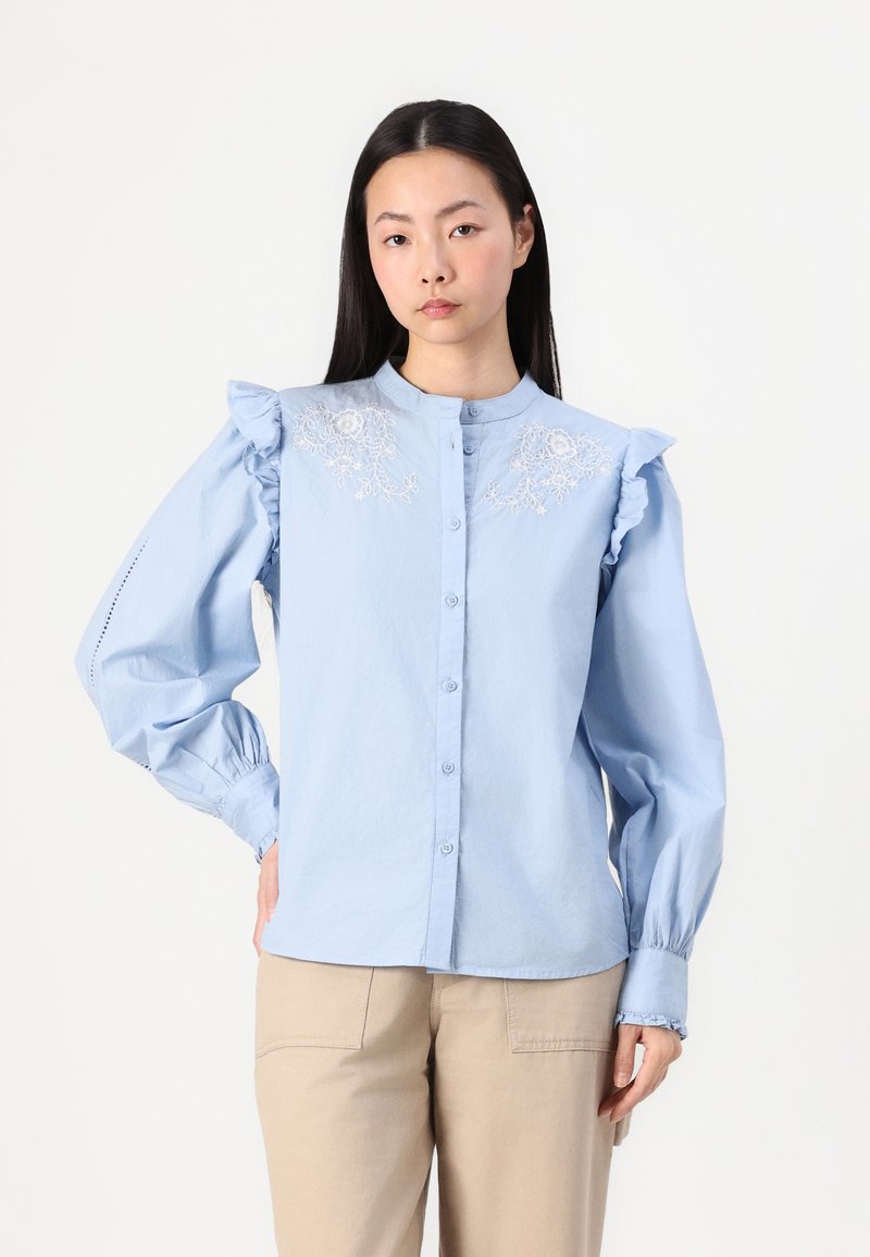 Blouse en coton bleu clair avec des accents de volants aux épaules, fermeture à boutons et broderie florale sur la poitrine. Manches longues et bouffantes avec des poignets froncés.
