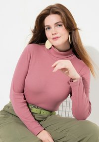 Helles rosa Langarm-Turtleneck-Oberteil mit geripptem Muster, kombiniert mit olivgrünen Hosen und einem passenden Gürtel; Model trägt große geflochtene Ohrringe.