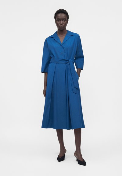 Γυναίκα με μεσαίου μήκους μπλε κοβαλτίου φόρεμα με ζώνη, μανίκια μέχρι τον αγκώνα και μαύρα μυτερά παπούτσια με τακούνι, στέκεται μπροστά σε ένα απλό φόντο.