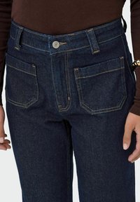 Jean en denim bleu foncé à taille haute, avec descoutures jaunes contrastantes, des poches avant et une fermeture à bouton en métal visible.