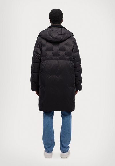 Geacă puffer neagră cu glugă, prezentând un design matlasat, mâneci lungi și croială dreaptă, asortată cu blugi de culoare bleu deschis și adidași albi.
