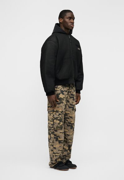 Μαύρο μπουφάν μάλλινο με κουκούλα, κλείσιμο με φερμουάρ και μπροστινές τσέπες, συνδυασμένο με μπεζ παντελόνια καμουφλάζ cargo και μαύρα αθλητικά παπούτσια.