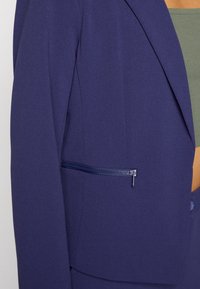 Námořnický modrý blazer s klopovým límcem a postranní kapsou na zip. Vyrobený z hladké tkaniny s strukturovaným střihem. Minimalistický design.