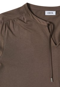 Haut en jersey brun avec un décolleté en V et des détails froncés aux épaules. Doté d'une fermeture à cordon avec des accents métalliques. Étiquette de la marque visible.