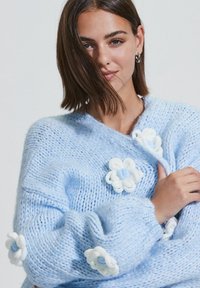 Modèle avec des cheveux mi-longs portant un pull en tricot bleu clair orné d'appliqués de fleurs blanches et bleues, souriant doucement.