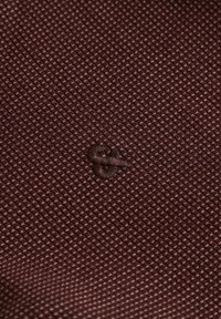 Gros plan sur un tissu texturé bordeaux avec de petits points blancs et un symbole esperluette cousu avec un fil bordeaux assorti.