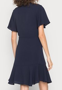 Tyrkysově modré krátké šaty s volány, měkká textil, vypasovaný pas a mnohoúrovňový volánový lem. Jednoduchý, elegantní design bez ozdob.