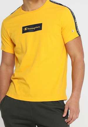 Mand iført en gul Champion t-shirt med sort logo på brystet og sorte mærkede striber på skuldrene, sat sammen med mørke bukser.