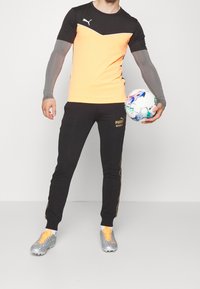 Černé a oranžové sportovní tričko se šedými rukávy, spárované s černými kalhotami se zlatými akcenty, držící multicolor fotbalový míč.