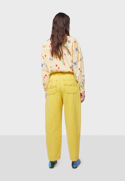 Pantalones amarillos de talle alto con piernas anchas, que presentan bolsillos bordados y una tela texturizada. Camisa de patrones multicolores, de mangas largas.