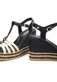 Sandalias de cuña negras con tiras de crema, detalles de yute trenzado y un empeine texturizado. Cuentan con correas ajustables en el tobillo y acentos en tono dorado.