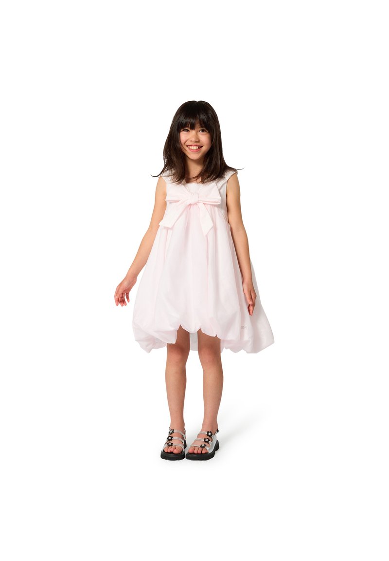 Φόρεμα ανοιχτό ροζ χωρίς μανίκια με μεγάλο φιόγκο στη μέση, ημιδιαφανές ύφασμα και στρογγυλεμένο τελείωμα. Συνδυάζεται με μαύρα σανδάλια με λουριά.