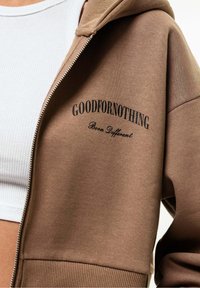 Hnědá mikina na zip s nápisem „GOODFORNOTHING“ a sloganem „Born Different“ v černé barvě. Má měkkou texturu a volný střih.