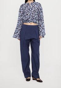 Blouse à manches longues imprimée de motifs floraux en nuances de bleu et de brun, nouée à la taille ; associée à un pantalon large marine et des ballerines noires.