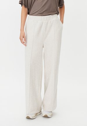 Pantalones sueltos de color beige claro con cintura elástica, combinados con una camiseta marrón de manga corta y zapatillas blancas sobre un fondo blanco.