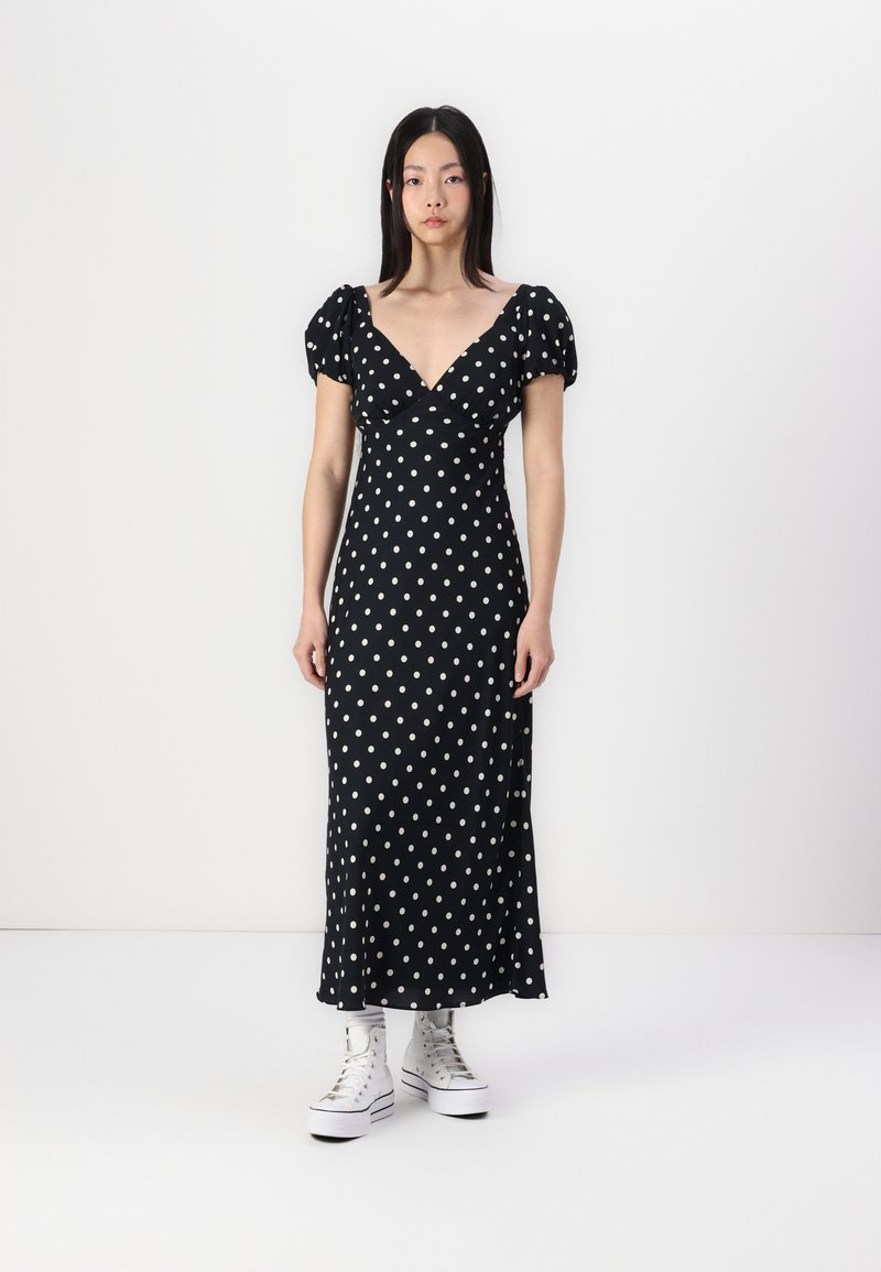 Robe noire à pois blancs, décolleté en V profond, manches courtes bouffantes et silhouette cintrée. Portée avec des baskets montantes blanches.