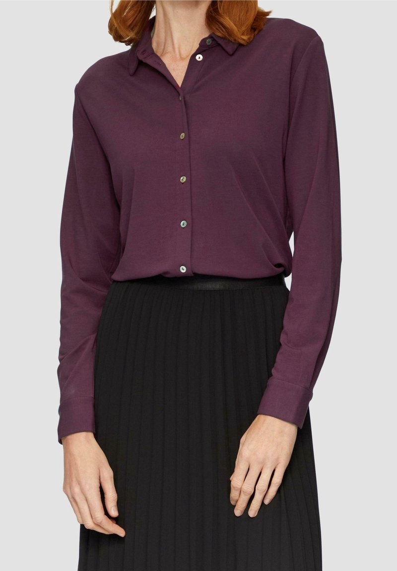 Femme portant une chemise violette à manches longues boutonnée, rentrée dans une jupe plissée noire taille haute, se tenant devant un fond uni.