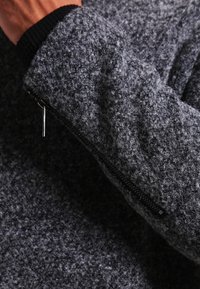 Jersey de lana gris con una superficie texturizada, que cuenta con un bolsillo con cremallera y puños acanalados. La tela parece gruesa y cálida.