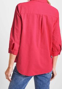 Chemise fuchsia à manches longues avec manches retroussables, dotée d'un détail plissé au dos et d'un ourlet arrondi, fabriquée en tissu léger.