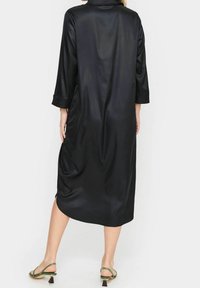 Svart satinklänning med krage, trekvartsärmar, sidokilar och en flödande silhuett. Matchas med gröna sandaler med remmar.