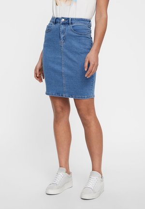 Kvinde iført en blå denim nederdel til knæene, hvide sneakers og en lysefarvet top, stående foran en ensfarvet baggrund.