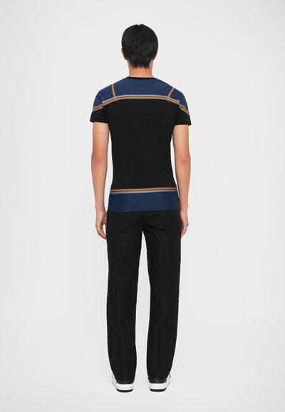 Camiseta negra con líneas de acento azules y naranjas en los hombros, combinada con pantalones negros texturizados y zapatillas blancas, vista desde atrás.