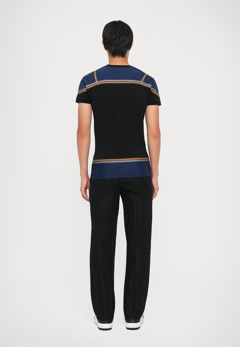 Zwarte t-shirt met blauwe en oranje accentlijnen op de schouders, gecombineerd met gestructureerde zwarte broek en witte sneakers, van achter bekeken.