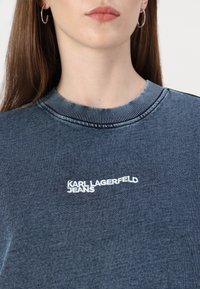 Temno modra pulover iz teksturirane tkanine, ki ima rebrast ovratnik in beli vezeni logotip "KARL LAGERFELD JEANS" na sprednji strani.