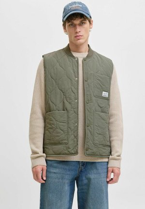 Jeune homme portant un gilet matelassé vert sur un pull beige, un jean bleu et une casquette bleue avec du texte blanc.