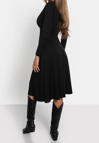 Femme aux longs cheveux portant une robe noire à manches longues et des bottes noires montant jusqu'aux genoux, debout de profil sur un fond uni.