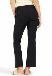 Pantalones acampanados negros con detalles de costura, confeccionados en tejido texturizado. Combinados con calzado tipo zueco en tono marrón, mostrando los pies descalzos.