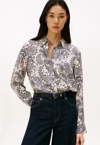 Vrouw die een crèmekleurig shirt met paisleypatroon en lange mouwen draagt, in donkerblauwe jeans met een zwarte riem, staand tegen een effen achtergrond.