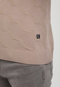 Beež pullover, millel on kuusnurga struktuuriline kudumine peene tekstuuriga ja soonikkoes äär. Väike must logo silt alumises vasakus servas.