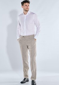 Mann in einem weißen Hemd mit Knöpfen, beigen Anzughosen und schwarzen Lederschuhen, steht mit den Händen in den Taschen und blickt nach links.