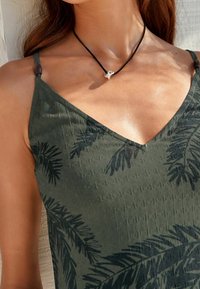 Robe vert olive à encolure en V, ornée de motifs de feuilles foncées, avec de fines bretelles et un tissu texturé. Collier en cordon noir avec un accent argenté.