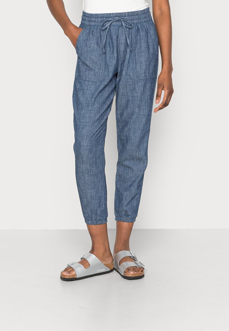 Pantalons jogger en denim bleu clair avec une taille élastique, un cordon de serrage, des poches latérales et des poignets de pantalon fuselés. Portés avec des sandales argentées.