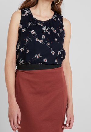 Femme portant un chemisier sans manches bleu marine à fleurs, rentré dans une jupe taille haute couleur rouille avec une ceinture noire, et des boucles d'oreilles créoles dorées.