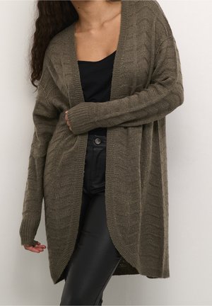 Kvinde iført en lang, brun, tekstureret cardigan over en sort top og sorte bukser, står op ad en ensfarvet lys væg.