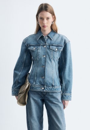 Jeune femme portant une veste en jean bleue ajustée et un jean, tenant une pochette tissée beige, debout devant un fond clair uni.