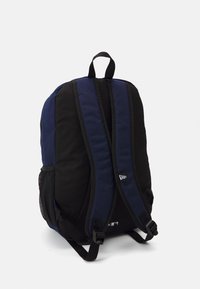 Sac à dos en tissu noir et marine avec des sangles rembourrées, une poignée en haut et des poches latérales en maille. Présente un logo dans la partie inférieure.