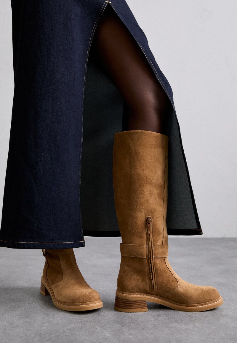 Des bottes en daim beige montantes avec un bout arrondi, un détail de fermeture éclair sur le côté et un talon empilé, assorties à une longue jupe en denim.