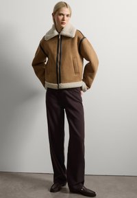 Chaqueta bomber de ante marrón con un cuello y puños de piel sintética blanca; cuenta con un cierre de cremallera negro y dos bolsillos frontales. Pantalones de pierna ancha marrón.