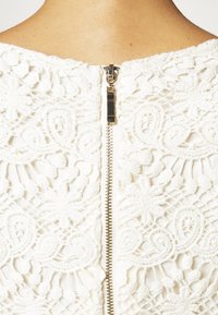 Vitt spetsklänning med intrikata blommönster, med en synlig metalldragkedja på baksidan. Spetsens struktur ger dimension och detaljer.