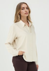 Beige Hemd mit Knöpfen, Kragen und langen Ärmeln, mit glatter Textur und abgerundetem Saum, getragen über braunen Hosen.